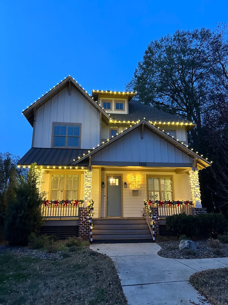 Two-story craftsman home with warm white roofline lights, wrapped porch columns, garland and red bows