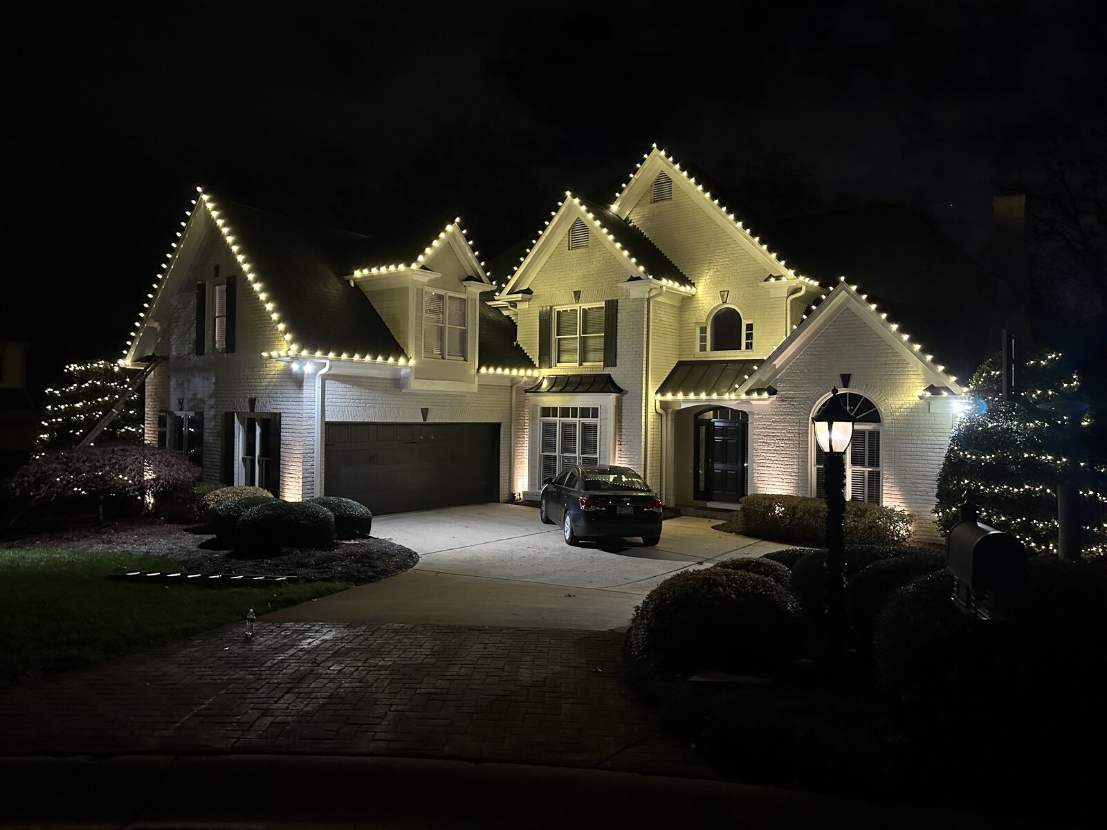 White brick home with warm white lights along every roofline and lit shrubs by the entry